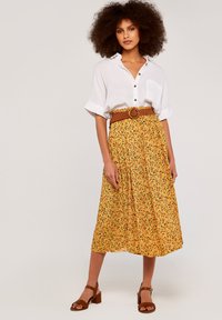 Gelber, blumiger Midirock mit Faltenwurf, kombiniert mit einem weißen Hemd mit Knöpfen und einem strukturierten braunen Gürtel. Braune Blockabsatzsandalen.