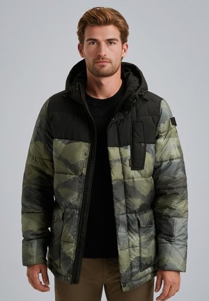 Homme portant une veste rembourrée à capuche en camouflage vert et noir, un shirt noir et un pantalon brun, se tenant devant un fond gris uni.