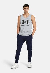 Šedé tielko bez rukávov s čiernym logom "Under Armour", skombinované s námorníckymi teplákmi a bielymi teniskami, ktoré predvádzajú uvoľnený atletický strih.