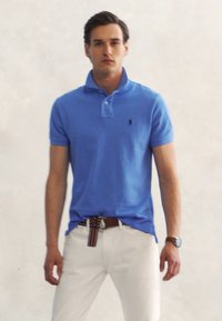 Άνδρας που φοράει μπλε πόλο πουκάμισο, μπεζ παντελόνι, ριγωτή ζώνη και ρολόι χειρός, στέκεται μπροστά σε φωτεινό ουδέτερο φόντο.