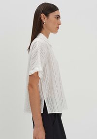 Camicia bianca a maniche corte con colletto, caratterizzata da un motivo intrecciato testurizzato e spacchi laterali. Indossata con pantaloni neri, che mettono in evidenza una vestibilità moderna.