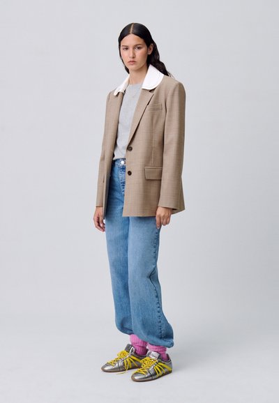 Blazer a quadri marrone con colletto bianco, camicia grigia, jeans azzurri chiari e sneakers argentate con accenti gialli e calzini rosa.