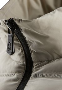 Beige puffjacka i tyg med nära vy av en dragkedja, som har en matt svart dragflik och texturerade sömmar längs kanterna.