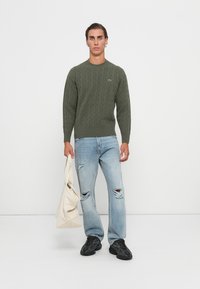 Olivengrønn kabelstrikket genser, tettsittende design, teksturert stoff. Brukt med lyseblå rippede jeans og svarte joggesko. Holder en beige totebag.