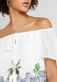 Blouse blanche à épaules dénudées avec des manches ondulées et un détail en tresse à l'avant. Elle présente un motif de paysage urbain et de palmiers au niveau de l'ourlet.