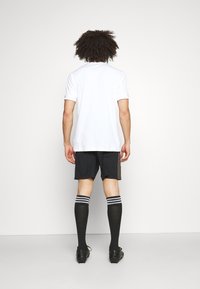 Witte sportshirt, zwarte shorts met zijstrepen, zwarte sokken met witte strepen en zwarte cleats. Achteraanzicht van de outfit.