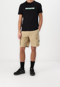 Schwarzes T-Shirt mit einem mintgrünen "Calvin Klein"-Logo, kombiniert mit beigen Cargo-Shorts mit Seitentaschen, getragen mit schwarzen Socken und Sneakers.