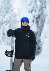 Snowboarder wearing a black jacket with brand logo, brown pants, and blue mirrored goggles, holding a snowboard against a snowy backdrop.