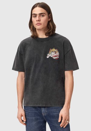 Młody mężczyzna z brązowymi włosami do ramion, ubrany w wyblakły, czarny, oversize'owy T-shirt z kolorowym logo "Allsaints Rock" oraz niebieskie dżinsy.