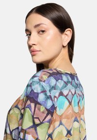 Bluzka w wielokolorowy wzór geometryczny wykonana z lekkiego, prześwitującego materiału. Okrągły dekolt i luźny fason, w odcieniach zieleni, niebieskiego i fioletu.