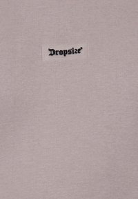 Tissu beige avec une petite étiquette blanche comportant le texte noir de style gothique « Dropsize » cousue sur la surface.