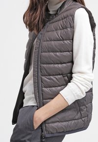 Gilet imbottito grigio scuro, con zip frontale e tasche laterali. Indossato sopra un dolcevita crema, mostra una texture trapuntata e una vestibilità moderna.