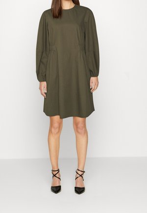 Robe en coton vert olive avec de longues manches bouffantes, côtés froncés et col rond. Portée avec des escarpins noirs à talons pointus agrémentés de lanières croisées.