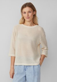 Maglione aperto beige chiaro con scollatura rotonda, maniche a tre quarti e vestibilità comoda. Indossato con pantaloni di jeans blu chiaro.