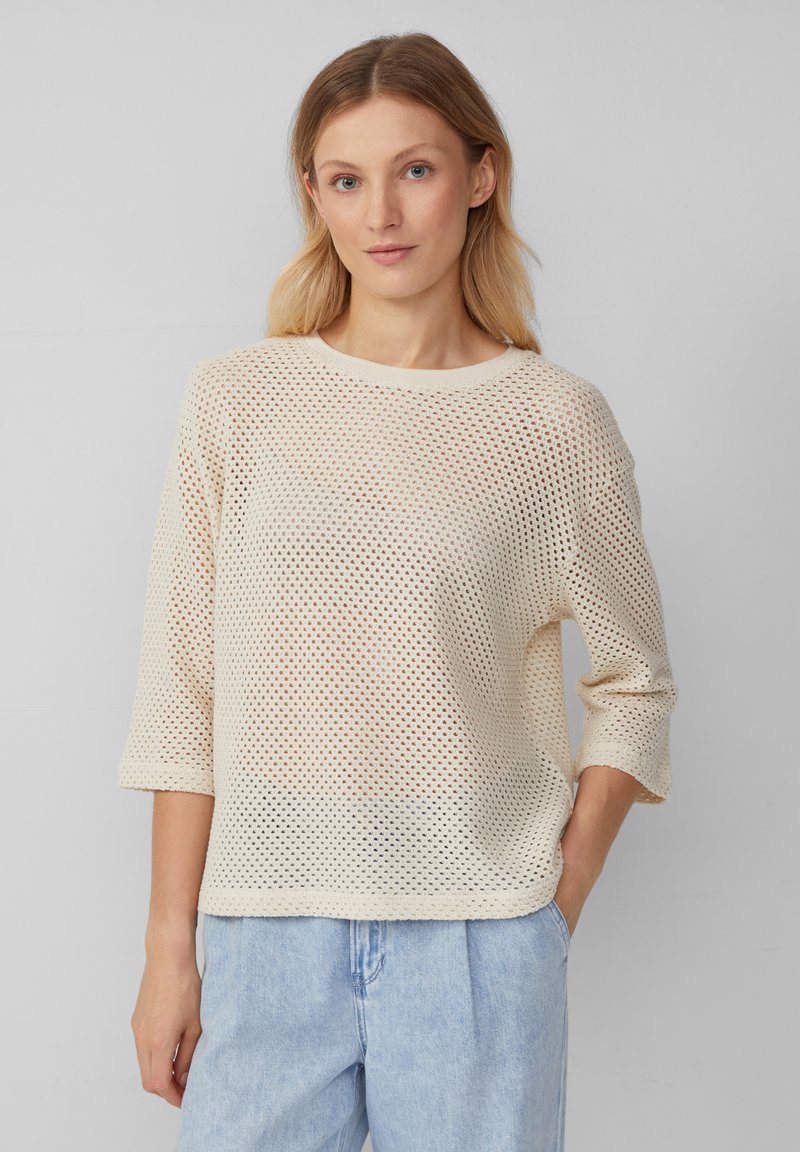 Maglione aperto beige chiaro con scollatura rotonda, maniche a tre quarti e vestibilità comoda. Indossato con pantaloni di jeans blu chiaro.