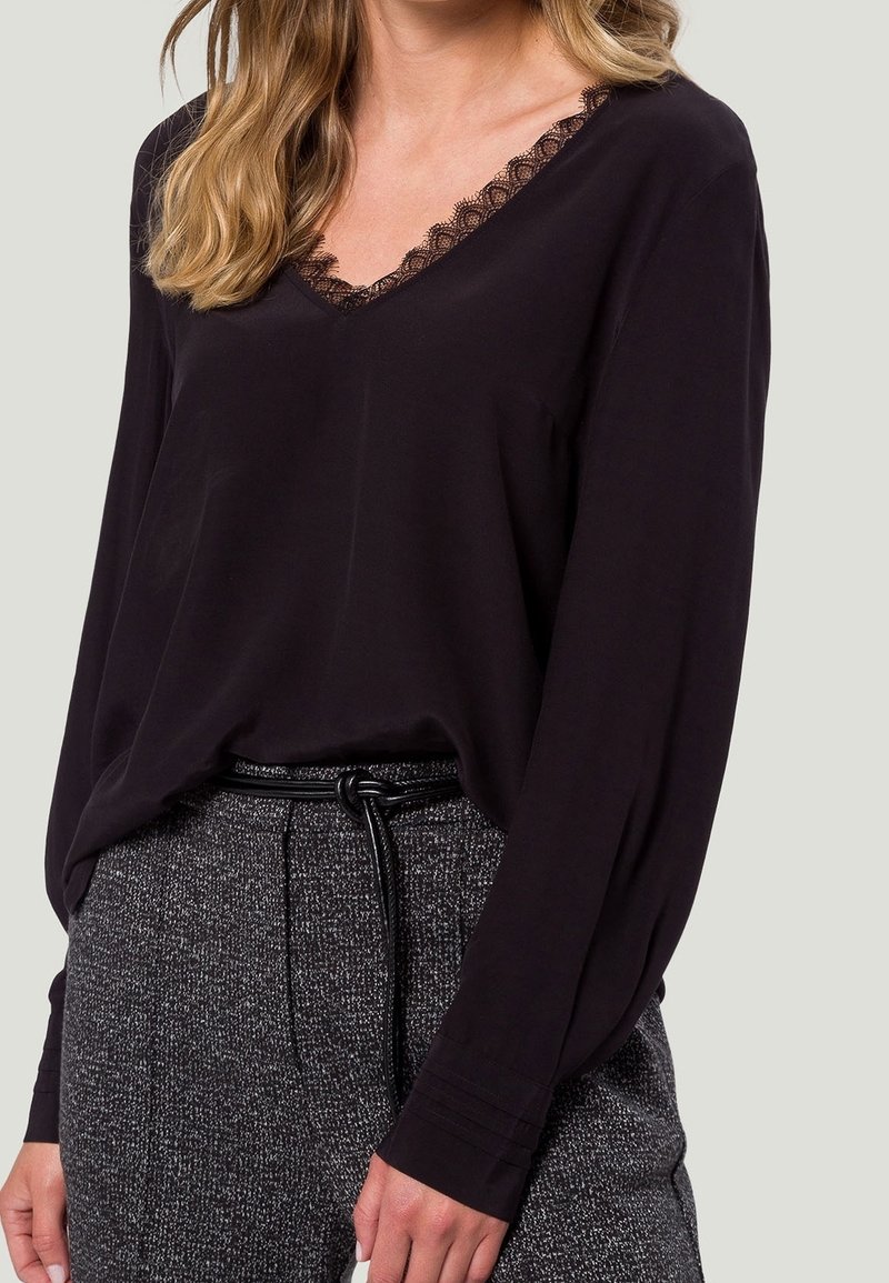 Femme portant un chemisier noir à manches longues en V avec un bord en dentelle, rentré dans un pantalon gris texturé taille haute avec une fine ceinture noire.