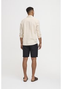 Maglietta beige a maniche lunghe, maniche arrotolate, abbinata a pantaloni corti neri. L'individuo indossa sandali scuri. Design semplice e casual senza motivi.