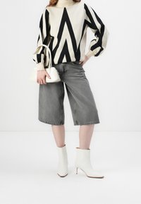 Personne portant un pull géométrique noir et blanc, un short gris large mi-genou, des bottines blanches à la cheville et tenant un petit sac à main blanc.