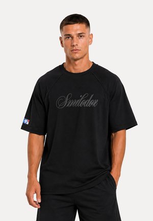 Joven con el pelo corto y oscuro que lleva una camiseta negra holgada con el texto "Smilodox" y pantalones negros, de pie sobre un fondo blanco.