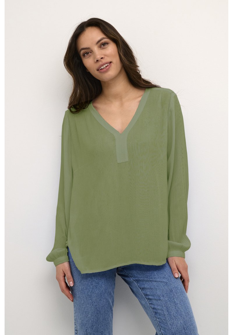 Vrouw met lang donker haar, gekleed in een losse lichtgroene V-hals blouse en blauwe jeans, staand tegen een witte achtergrond.
