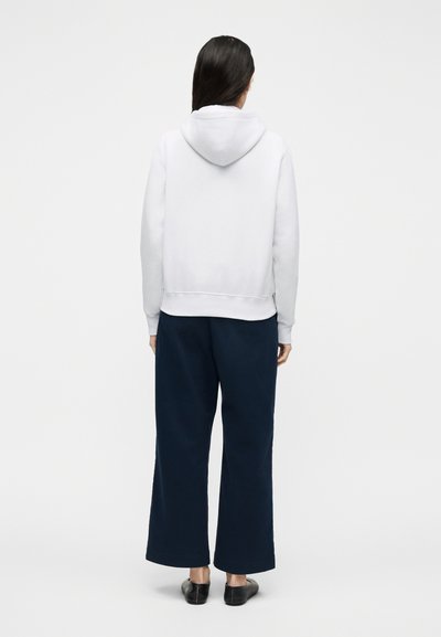 Persona con capelli lunghi e scuri che indossa una felpa con cappuccio bianca, pantaloni larghi blu navy e scarpe nere senza lacci, in piedi davanti a uno sfondo chiaro e uniforme.