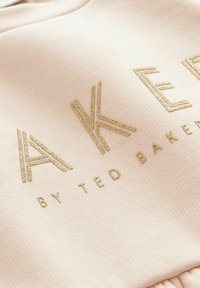 Felpa di tessuto rosa pallido con lettering metallico dorato che recita "AKER BY TED BAKER", con texture a coste e scollatura rotonda.