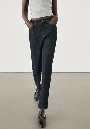 Personne portant un jean droit bleu foncé, un top court gris clair et des chaussures noires pointues avec des boucles, debout les mains dans les poches.