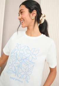 T-shirt en coton blanc avec un imprimé floral bleu clair et le texte "continuez à grandir". Encolure ronde et manches courtes.
