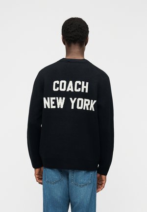 Person trägt einen schwarzen Pullover mit der Aufschrift "COACH NEW YORK" in weißen Buchstaben auf der Rückseite, kombiniert mit blauen Jeans, und steht mit dem Rücken zur Kamera.