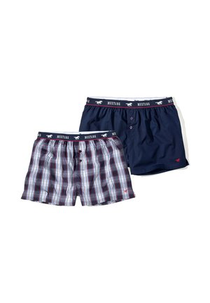 Lächelnder, oberkörperfreier Mann, der blaue, weiße und rote karierte Boxershorts mit "Mustang"-Logo am Bund vor einfarbigem Hintergrund trägt.