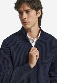 Pull en maille côtelée marine avec un col haut et une fermeture éclair partielle. Présente un tissu texturé et une silhouette ajustée, porté sur une chemise à col.