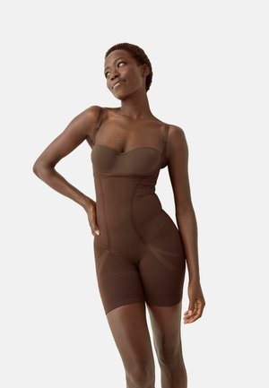 Kobieta w brązowym modelującym body, stoi z jedną ręką na biodrze i patrzy w bok, jednolite jasne tło.
