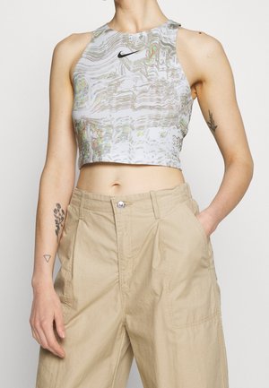 Ärmelloses Crop-Top mit verzerrtem Marmor-Muster und schwarzem Nike-Logo, getragen zu hochtaillierten beigen Hosen, zeigt Unterarm-Tattoos und Hand in der Tasche.
