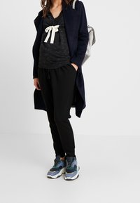 Svart långärmad topp med krämfärgad rosett, tillsammans med svarta joggers och färgglada sneakers. Marinblå kappa hänger över axlarna. Ryggsäck på ryggen.