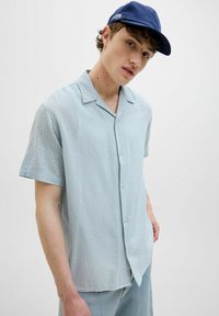Camisa de mangas cortas de color azul claro con un patrón a cuadros, con botón en la parte delantera y un corte relajado. Combinada con jeans de mezclilla claros para un look casual.