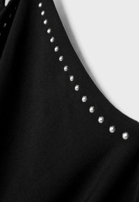 Czarny materiał o gładkiej fakturze, z rzędem małych srebrnych ćwieków wzdłuż dekoltu, podkreślający detale wzoru.