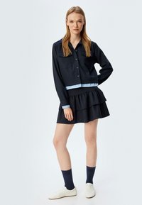 Giacca accorciata a righe navy con profili blu e maniche lunghe, abbinata a una gonna nera a strati. Sneakers bianche e calzini scuri completano il look.