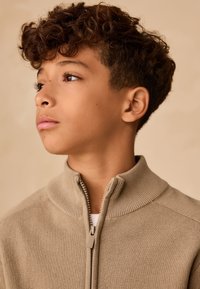 Maglione beige con zip e collo alto, con texture a coste. Il modello ha capelli corti, ricci e castani, e indossa una camicia bianca sotto.