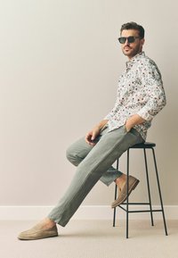 Chemise à boutons florale blanche, pantalon en lin gris, mocassins beiges et lunettes de soleil noires. Modèle assis sur un tabouret noir contre un fond uni.