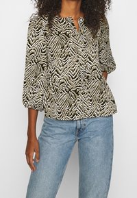 Blusa verde oliva con patrón de zigzag blanco, escote redondo, cierre de botón y mangas abullonadas de tres cuartos, combinada con jeans de color azul claro.