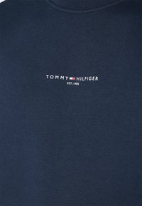 T-shirt in cotone blu navy con collo a girocollo a costine, caratterizzato dal logo bianco "TOMMY HILFIGER" e "EST. 1985" in un bold sans-serif.