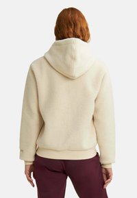 Felpa color crema con superficie texture, cappuccio con coulisse e polsini a costine. Abbinata a pantaloni di un bordeaux scuro.