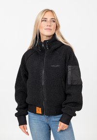 Veste en polaire noire avec une fermeture éclair sur le devant, un col haut et des poignets côtelés. Elle dispose d'une poche sur le bras gauche et d'une étiquette de marque orange au niveau de la taille.