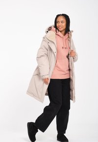 Lang beige pufferjakke med pelsforing i hætten, båret over en pink hættetrøje og sorte bukser. Jakken har elastiske manchetter og lynlås lukning.