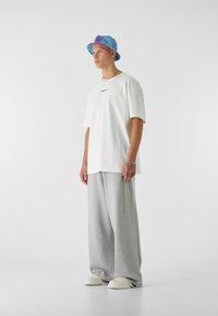 Biely tričko s malým čiernym logom, v kombinácii s voľnými šedými teplákmi a farebným batikovým klobúkom. Biely obuv dotvára outfit.