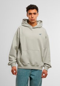 Hoodie vaaleanharmaana, pehmeästä kankaasta valmistettu, jossa on kenguru-tasku ja brodeerattu logo rinnassa. Pitkät hihat ja rento istuvuus.