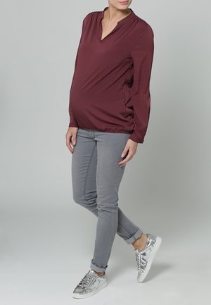 Haut de maternité en tissu bordeaux avec une coupe décontractée, un col en V et des manches longues, assorti à un jean gris slim et des baskets argentées.