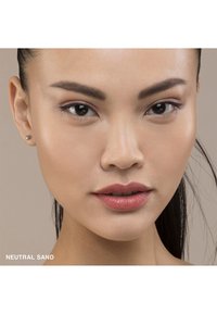 Nært bilde av et ansikt med glatt hud, medium brune toner, subtil sminke, nøytrale sandfargede lepper, perlestiftøreringer og mørkt hår trukket tilbake.