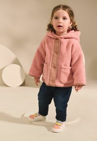 Puffige rosa Fleecejacke mit Kapuze und beiger Einfassung, mit zwei vorderen Taschen. Tragen mit dunklen Jeans und mehrfarbigen Sneakern.