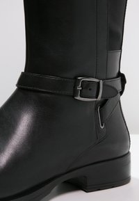 Bottine en cuir noir avec une sangle décorative et une boucle argentée autour du bas du mollet, détails cousus et petit talon bloc.